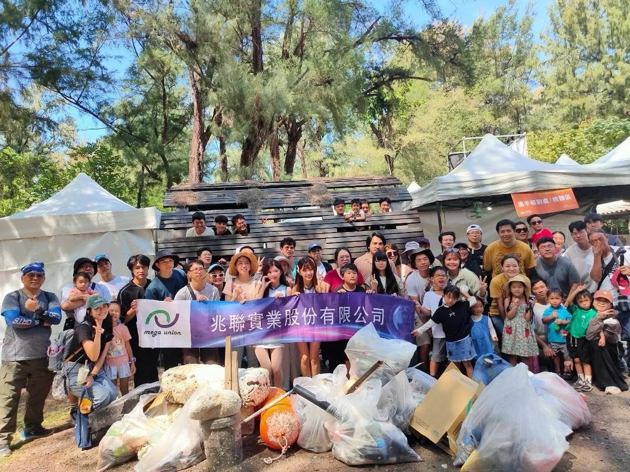 參與團隊在漁光島樹林旁拉著企業布條合照，前方擺放多袋淨灘收集物